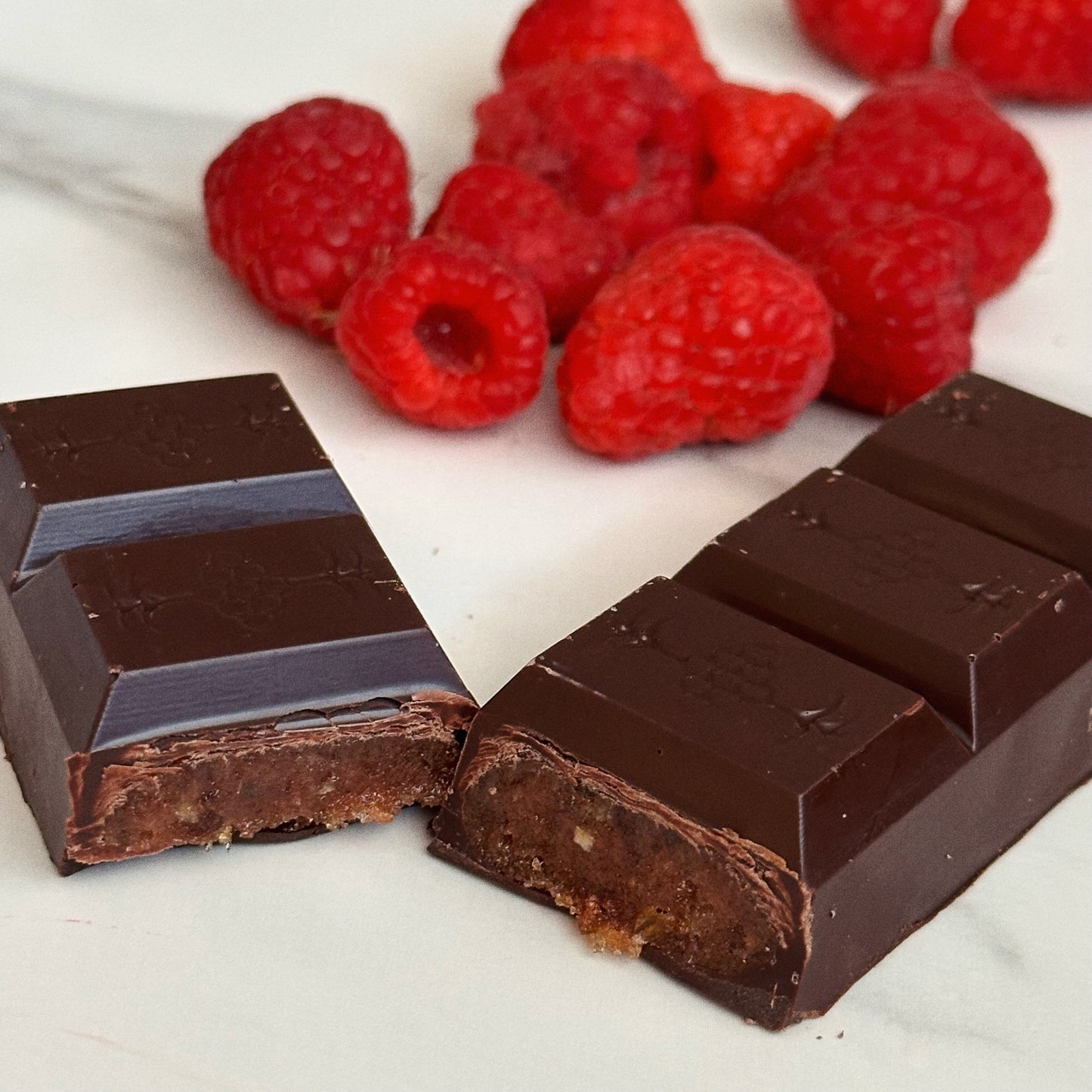 Dark chocolate bars with a piece broken off, surrounded by raspberries on a white surface.