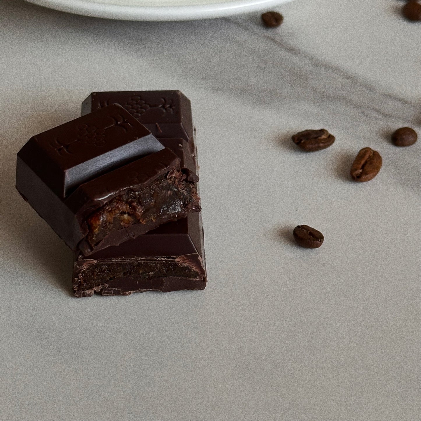 Dark chocolate bar with a piece broken off, coffee beans on a marble surface. Chewy Filling.